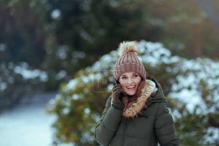 Téléchargez les photos : Femme d'âge moyen moderne souriante en manteau vert et chapeau brun à l'extérieur dans le parc de la ville en hiver avec des mitaines et un bonnet près des branches enneigées parlant sur un smartphone. - en image libre de droit