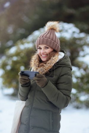 Photo for Happy modern middle aged woman in green coat and brown hat outdoors in the city park in winter with mittens and beanie hat sending text message using smartphone. - Royalty Free Image