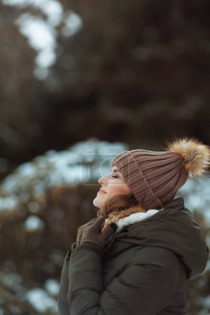 Téléchargez les photos : Femme d'âge moyen moderne souriante en manteau vert et chapeau brun à l'extérieur dans le parc de la ville en hiver avec des mitaines et un bonnet. - en image libre de droit