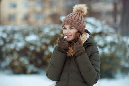 heureuse femme d'âge moyen moderne en manteau vert et chapeau brun à l'extérieur dans le parc de la ville en hiver avec mitaines, rouge à lèvres hygiénique et bonnet près des branches enneigées.