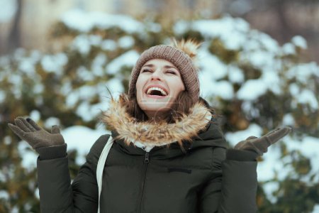 Téléchargez les photos : Femme moderne souriante en manteau vert et chapeau brun à l'extérieur dans le parc de la ville en hiver avec des mitaines et bonnet chapeau près des branches enneigées. - en image libre de droit
