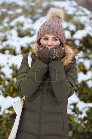 Téléchargez les photos : Heureuse femme d'âge moyen moderne en manteau vert et chapeau brun à l'extérieur dans le parc de la ville en hiver avec des mitaines et bonnet chapeau chaud mains froides avec souffle près des branches enneigées. - en image libre de droit