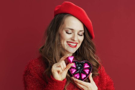 Téléchargez les photos : Joyeux Valentin. sourire élégant femme de 40 ans en pull rouge et béret avec boîte à bonbons en forme de coeur. - en image libre de droit