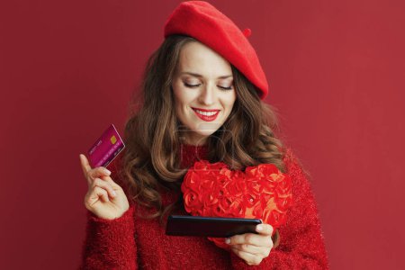 Téléchargez les photos : Joyeux Valentin. chère femme élégante de 40 ans en pull rouge et béret avec coeur rouge, smartphone et carte de crédit acheter en ligne. - en image libre de droit