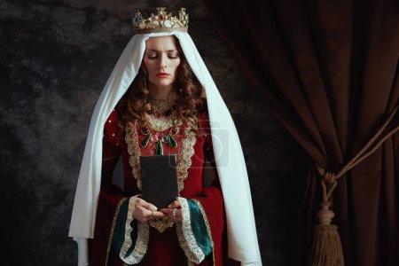 Photo for Medieval queen in red dress with book, veil and crown on dark gray background. - Royalty Free Image