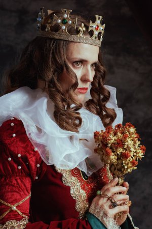 Foto de Triste reina medieval en vestido rojo con flor seca, cuello blanco y corona sobre fondo gris oscuro. - Imagen libre de derechos