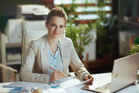 Téléchargez les photos : Portrait de femme comptable moderne heureuse en costume d'affaires léger dans un bureau vert moderne avec des documents et un ordinateur portable. - en image libre de droit