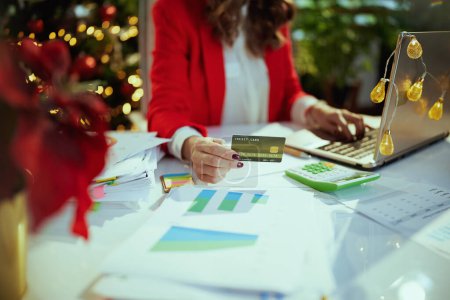 Photo for Christmas time. Closeup on middle aged business woman in santa hat and red jacket with documents, laptop and credit card working in green office with Christmas tree. - Royalty Free Image