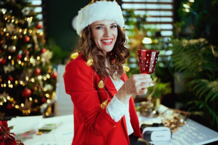 Téléchargez les photos : Noël. heureux moderne en veste rouge avec verre à vin en santa chapeau dans le bureau vert moderne avec arbre de Noël. - en image libre de droit