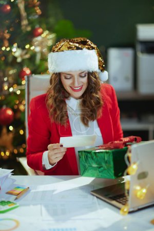 Photo for Christmas time. smiling modern in red jacket with present box and postcard in santa hat in modern green office with Christmas tree. - Royalty Free Image