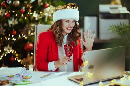 Téléchargez les photos : Noël. souriant élégant dans une veste rouge avec ordinateur portable et verre à vin en utilisant le chat vidéo dans le chapeau de Père Noël dans le bureau vert moderne avec arbre de Noël. - en image libre de droit