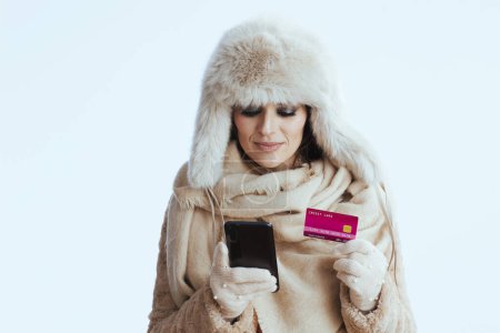 Photo for Stylish middle age woman in winter coat and fur hat against white background in white gloves with credit card and smartphone. - Royalty Free Image