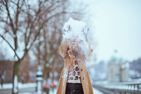 Téléchargez les photos : Femme souriante élégante de 40 ans en chapeau marron et écharpe en manteau de chameau jetant de la neige à l'extérieur en ville en hiver. - en image libre de droit