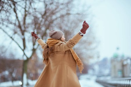 Téléchargez les photos : Vu de derrière femelle en chapeau marron et écharpe en manteau de chameau avec des gants se réjouissant à l'extérieur dans la ville en hiver. - en image libre de droit