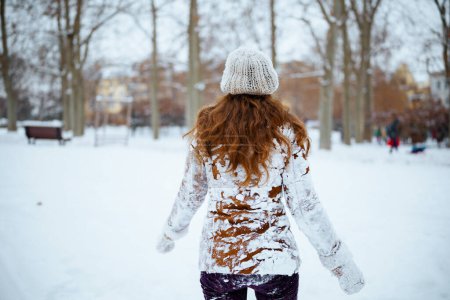 Photo for Seen from behind woman in brown hat and scarf in snowy clothes and sheepskin coat with mittens outside in the city in winter. - Royalty Free Image