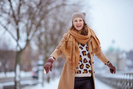 Foto de Elegante hembra sonriente en sombrero marrón y bufanda en abrigo de camello con guantes afuera en la ciudad en invierno. - Imagen libre de derechos