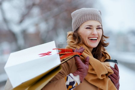 Téléchargez les photos : Femme élégante souriante en chapeau marron et écharpe en manteau de chameau avec gants, sacs à provisions et tasse de thé à l'extérieur de la ville en hiver. - en image libre de droit