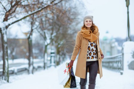 Foto de Feliz mujer moderna de 40 años en sombrero marrón y bufanda en abrigo de camello con guantes y bolsas de compras afuera en la ciudad en invierno. - Imagen libre de derechos