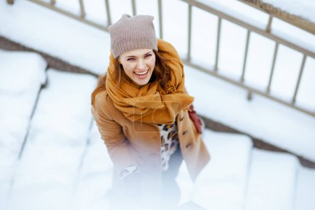 Photo for Upper view of smiling modern female in brown hat and scarf in camel coat with shopping bags outside in the city in winter. - Royalty Free Image