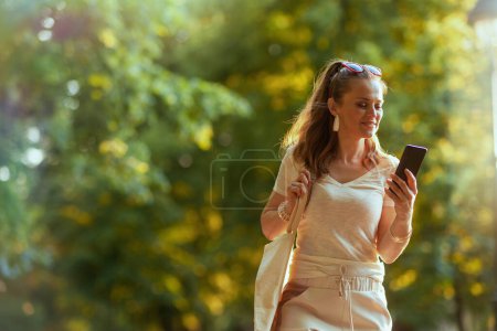 Foto de Hora de verano. mujer elegante pensativa en camisa blanca con teléfono inteligente y bolso de mano caminando en el parque de la ciudad. - Imagen libre de derechos