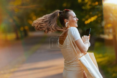 Téléchargez les photos : L'heure d'été. Femme souriante et tendance en chemise blanche avec smartphone et sac fourre-tout marchant dans le parc de la ville. - en image libre de droit