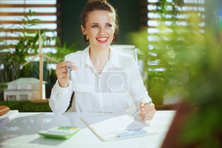 Photo for Eco real estate business. happy elegant 40 years old woman realtor in modern green office in white blouse with calculator, clipboard, document and keys. - Royalty Free Image