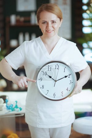 Photo for Healthcare time. happy massage therapist woman in spa salon with clock. - Royalty Free Image