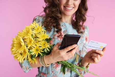 Photo for Closeup on smiling elegant woman with long wavy brunette hair with yellow chrysanthemums flowers and credit card using smartphone applications isolated on pink. - Royalty Free Image