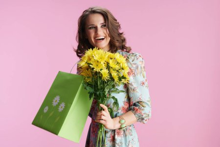 Photo for Happy trendy female with long wavy brunette hair with yellow chrysanthemums flowers and green shopping bag against pink background. - Royalty Free Image