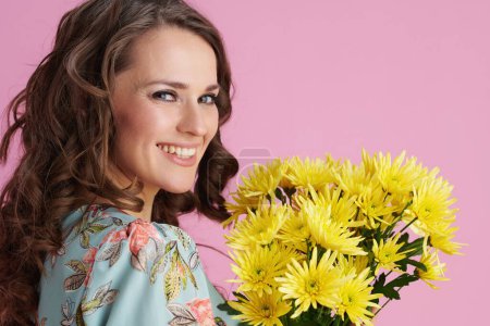 Foto de Retrato de mujer moderna sonriente con el pelo largo y ondulado morena con flores de crisantemos amarillos sobre fondo rosa. - Imagen libre de derechos