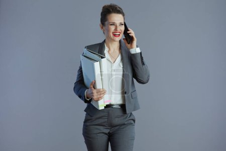 Foto de Sonriendo 40 años mujer trabajadora en traje gris con carpetas hablando en un teléfono celular contra fondo gris. - Imagen libre de derechos