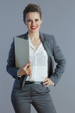 Foto de Sonriente mujer de negocios moderna de mediana edad en traje gris con portapapeles aislado sobre fondo gris. - Imagen libre de derechos