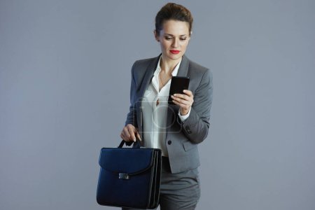 Foto de Mujer elegante y pensativa propietaria de una pequeña empresa en traje gris con teléfono inteligente y maletín aislado en gris. - Imagen libre de derechos