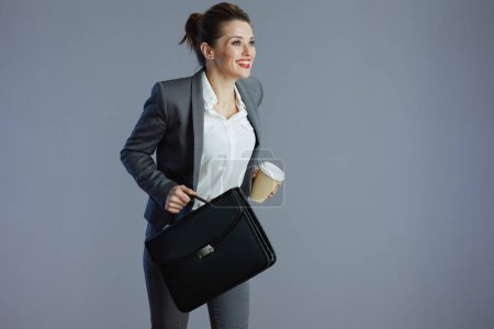 Foto de Sonriente empleada femenina elegante en traje gris con taza de café y maletín corriendo aislado sobre fondo gris. - Imagen libre de derechos