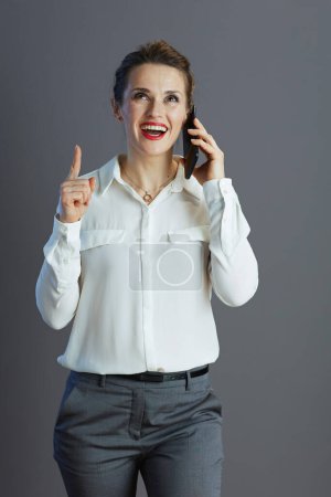 Téléchargez les photos : Heureuse élégante employée en chemisier blanc pointant vers le haut et parlant sur un smartphone isolé sur gris. - en image libre de droit