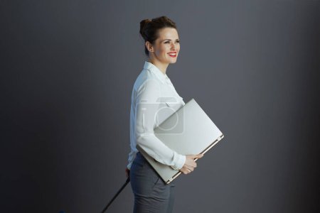 Foto de Sonriente elegante mujer de mediana edad trabajadora en blusa blanca con portátil y bolsa de ruedas aislada en gris. - Imagen libre de derechos