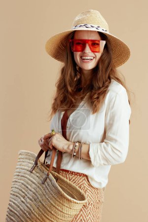 Foto de Vacaciones en playa. mujer moderna feliz en blusa blanca y pantalones cortos contra fondo beige con bolsa de paja, sombrero de verano y gafas de sol. - Imagen libre de derechos