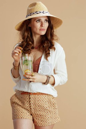 Photo for Beach vacation. trendy middle aged woman in white blouse and shorts isolated on beige background with cocktail and straw hat. - Royalty Free Image
