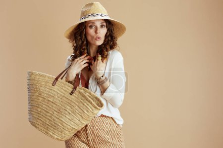 Photo for Beach vacation. surprised elegant middle aged housewife in white blouse and shorts isolated on beige background with straw bag and straw hat. - Royalty Free Image