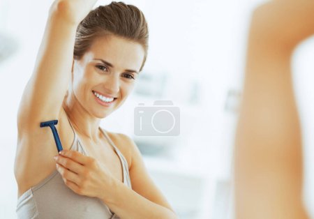 Téléchargez les photos : Heureuse jeune femme utilisant aisselle rasage dans la salle de bain - en image libre de droit