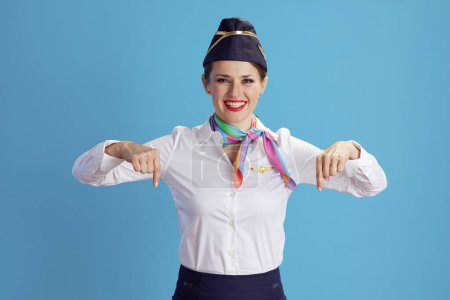 Téléchargez les photos : Souriante hôtesse de l'air féminine moderne sur fond bleu en uniforme pointant vers l'espace de copie. - en image libre de droit