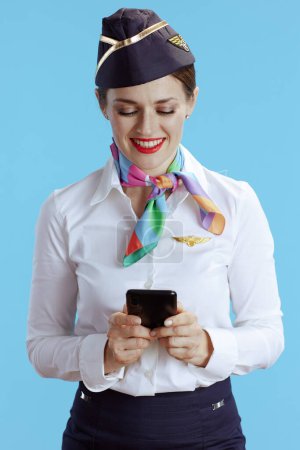 Photo for Smiling elegant female flight attendant against blue background in uniform using smartphone applications. - Royalty Free Image