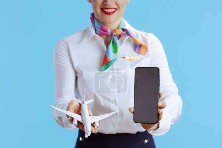 Foto de Primer plano de la azafata elegante aire femenino sobre fondo azul en uniforme que muestra la pantalla en blanco del teléfono inteligente. - Imagen libre de derechos