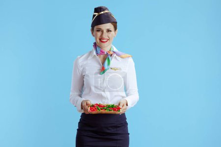 Photo for Smiling stylish female flight attendant isolated on blue background in uniform with candies. - Royalty Free Image
