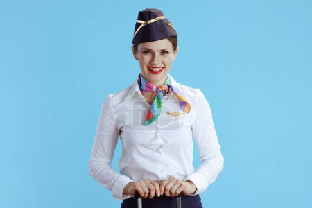 Foto de Feliz mujer auxiliar de vuelo moderno sobre fondo azul en uniforme con bolsa de ruedas. - Imagen libre de derechos