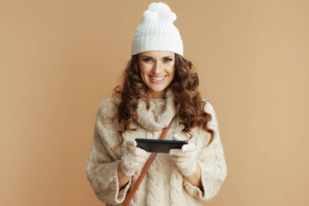 Photo for Hello winter. Portrait of happy modern 40 years old woman in beige sweater, mittens and hat using smartphone app isolated on beige. - Royalty Free Image