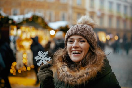 Foto de Joven hembra sonriente con abrigo verde y sombrero marrón en la feria de invierno en la ciudad con copo de nieve. - Imagen libre de derechos
