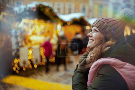 Téléchargez les photos : Heureuse femme d'âge moyen moderne en manteau vert et chapeau brun à la foire de Noël dans la ville en regardant au loin. - en image libre de droit