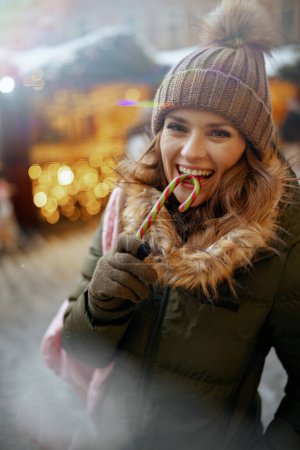 Téléchargez les photos : Femme moderne souriante en manteau vert et chapeau brun à la foire d'hiver dans la ville avec canne à bonbons. - en image libre de droit