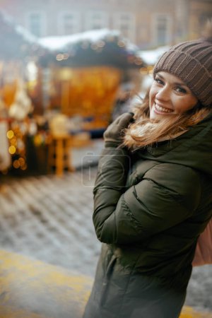 Foto de Sonriente mujer moderna de 40 años con abrigo verde y sombrero marrón en la feria de invierno en la ciudad. - Imagen libre de derechos
